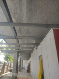 Structure métallique d'un porche de construction. Poutres grises sous un plafond texturé. Mur en panneaux isolants préfabriqués.