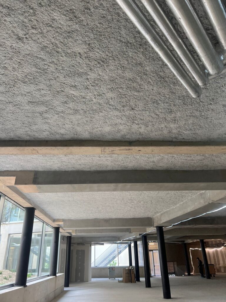 Flocage acoustique brut sur le plafond en béton d'un bâtiment moderne en construction, avec des poutres apparentes et des poteaux noirs cylindriques.